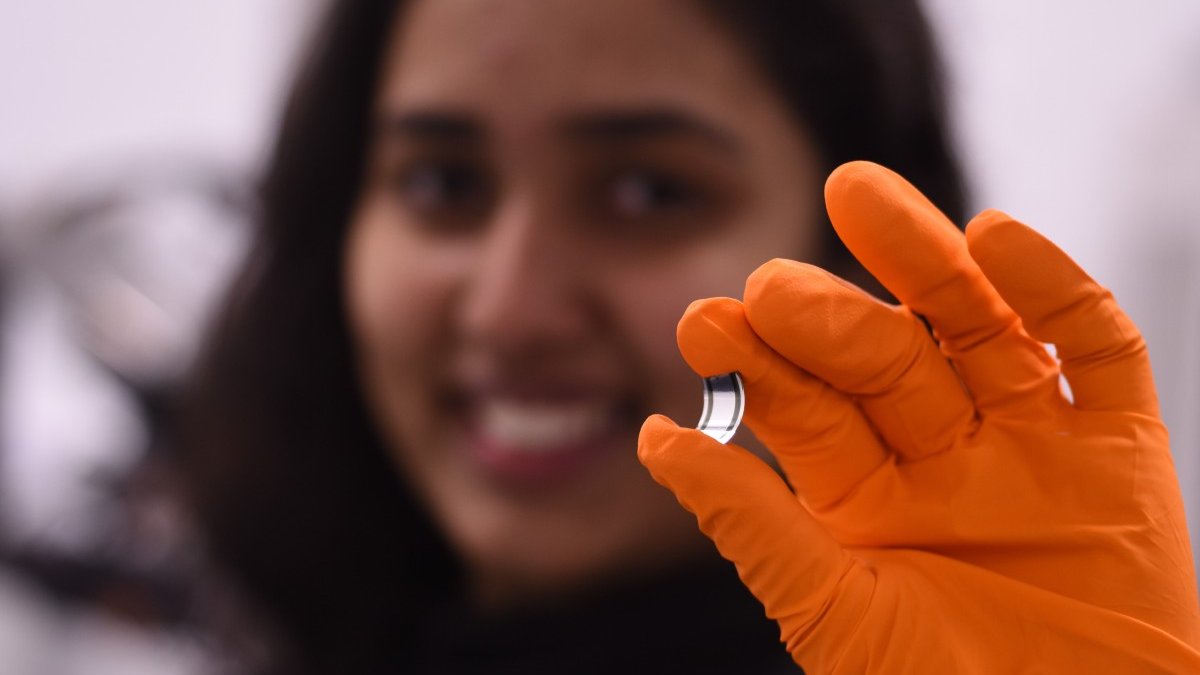 A blurred face smiles behind a hand, clad in an orange lab glove, holds a tiny rectangle of the new x-ray