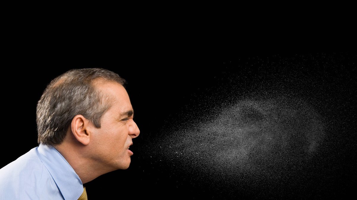 A middle aged white man wearing shirt and tie coughs. Against the plain black background, the spray from his mouth can be seen