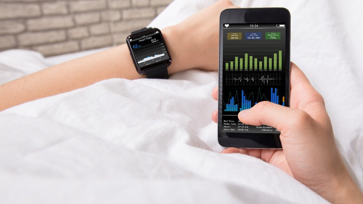 Two hands of a person lying in a bed with crisp white sheets. The left wrist wears a smart watch showing health data. The right hand holds a mobile phone
