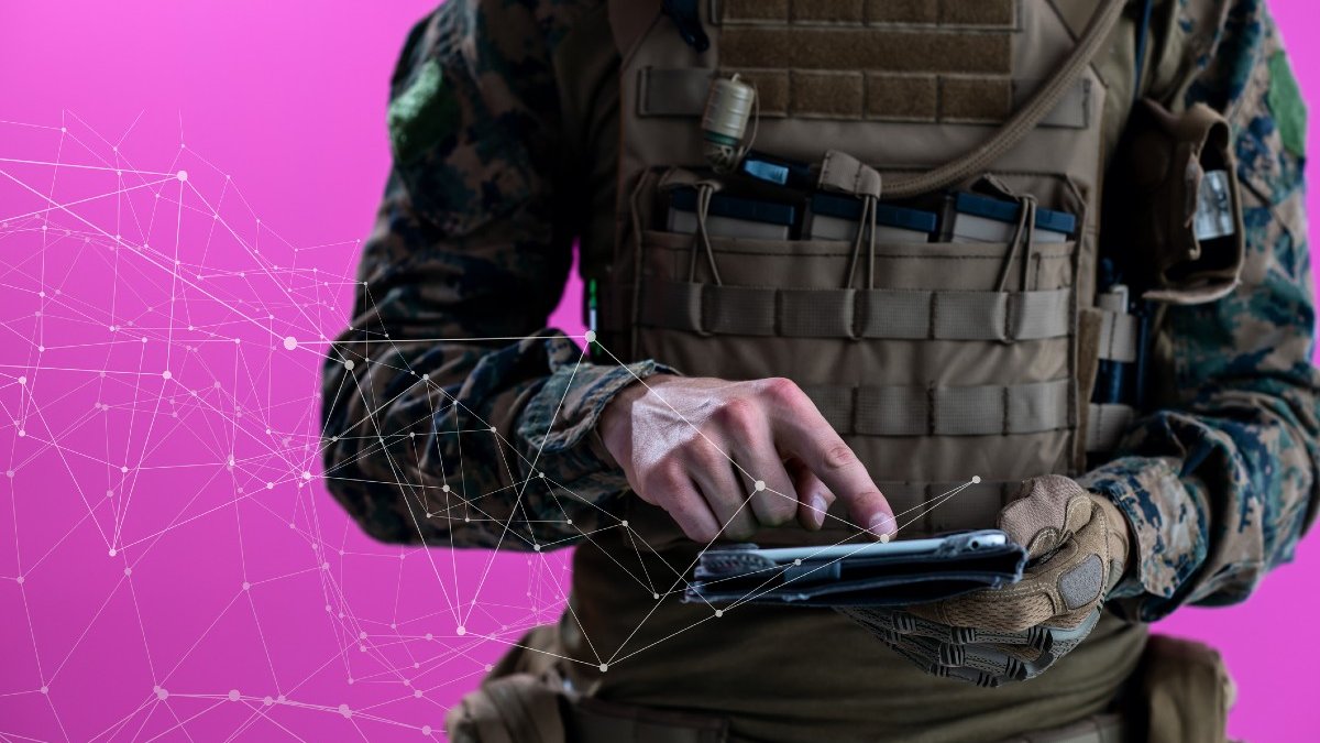 A soldier in combat uniform uses a tablet computer in front of a pink background with network concept lines