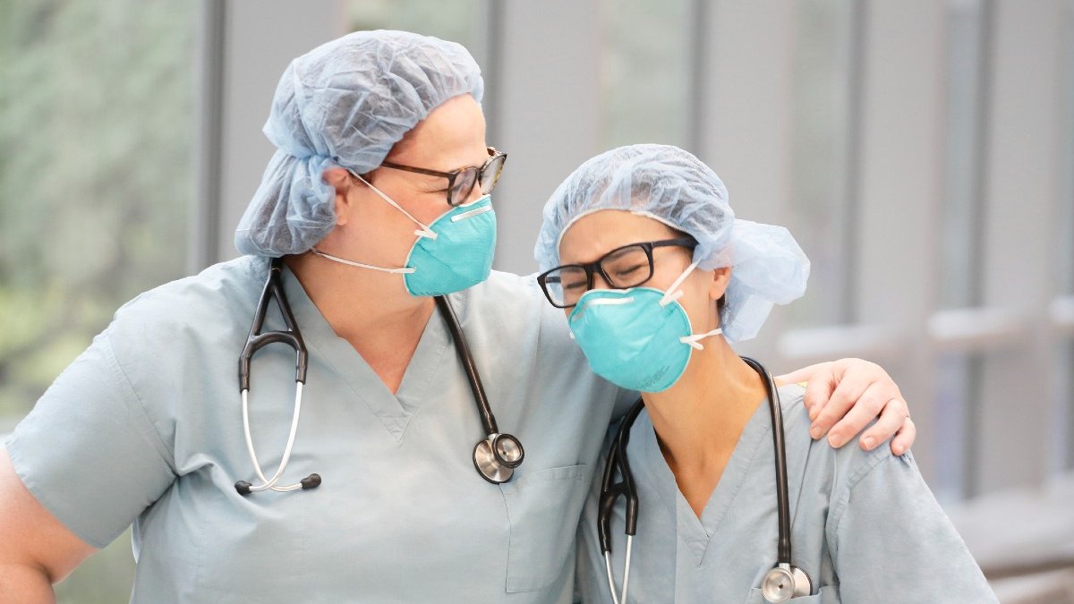 Two female, health care providers embrace while wearing light green medical scrubs, stethoscopes and N95 masks in an indoor hallway