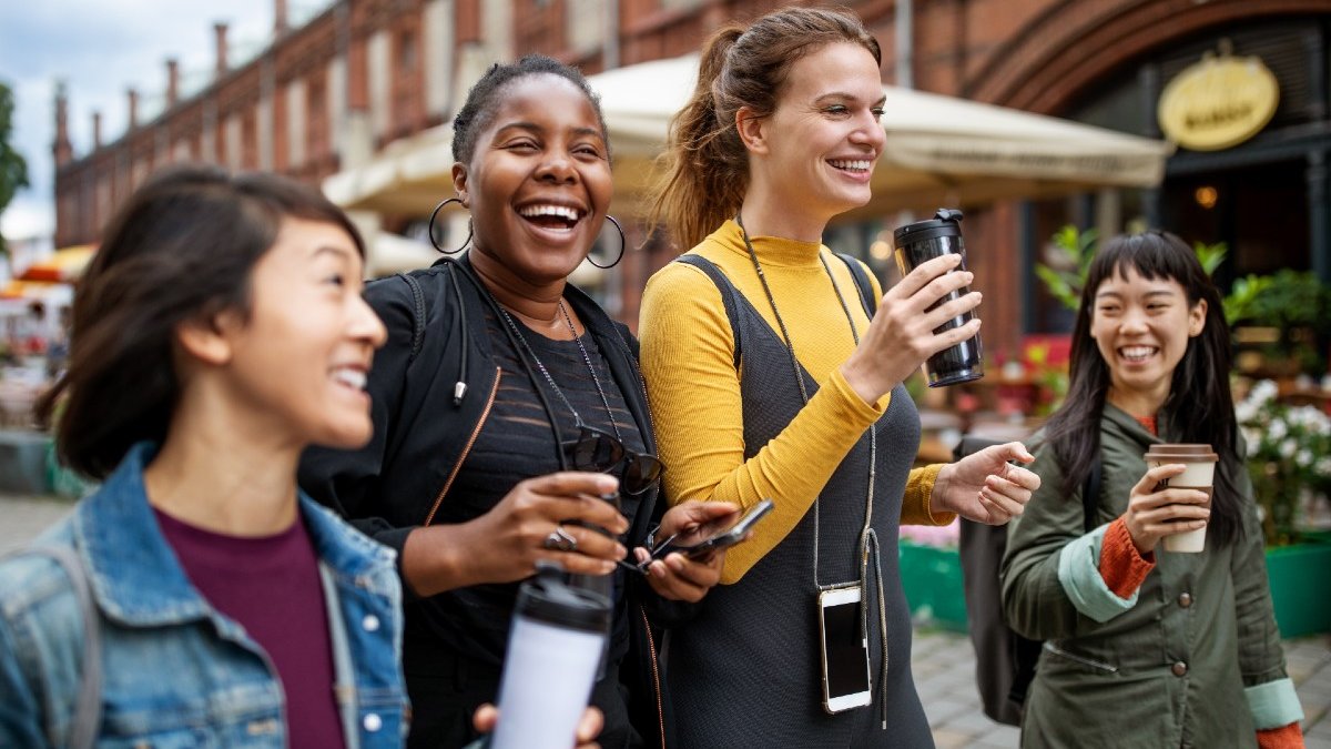 Four young women of different ethnic backgrounds enjoy a city break together. They are carrying phones and hot drinks and look as though they are having fun