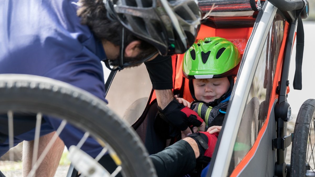 A dad wearing a silver bike helmet adjusts the straps of a toddler wearing a green bike helmet as he sits in a bike trailer