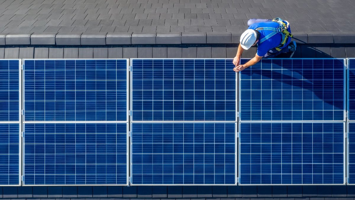 An aerial shot of a person wearing a helmet and blue top working on solar panels on a roof