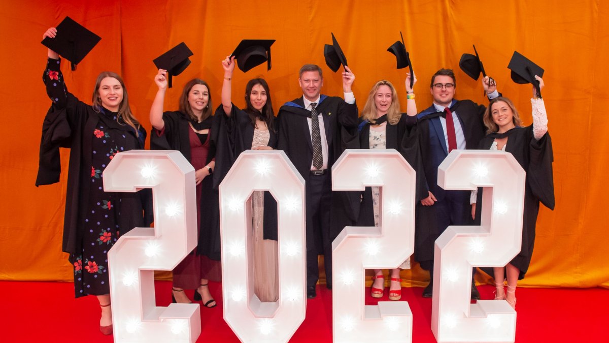 A photo of a group of graduates standing behind a lit-up 2022 sign, holding their caps in the air