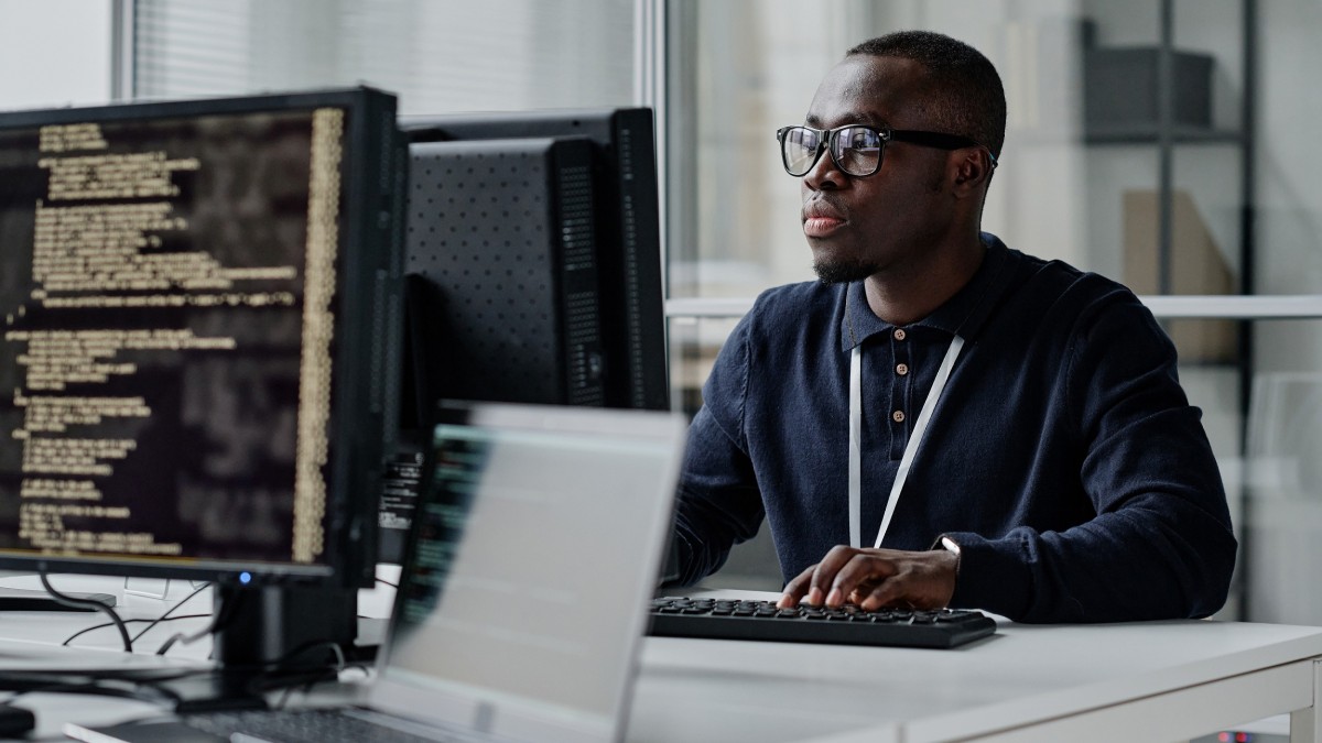 African American young developer in eyeglasses concentrating on his online work on computer sitting at workplace