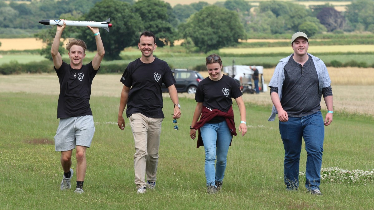 Four members of the Peryton Space team walk across a field, one holding their small rocket above his head