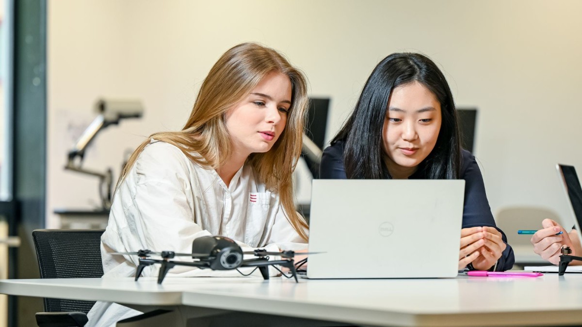 Two young women work together on a computer with a drone alongside