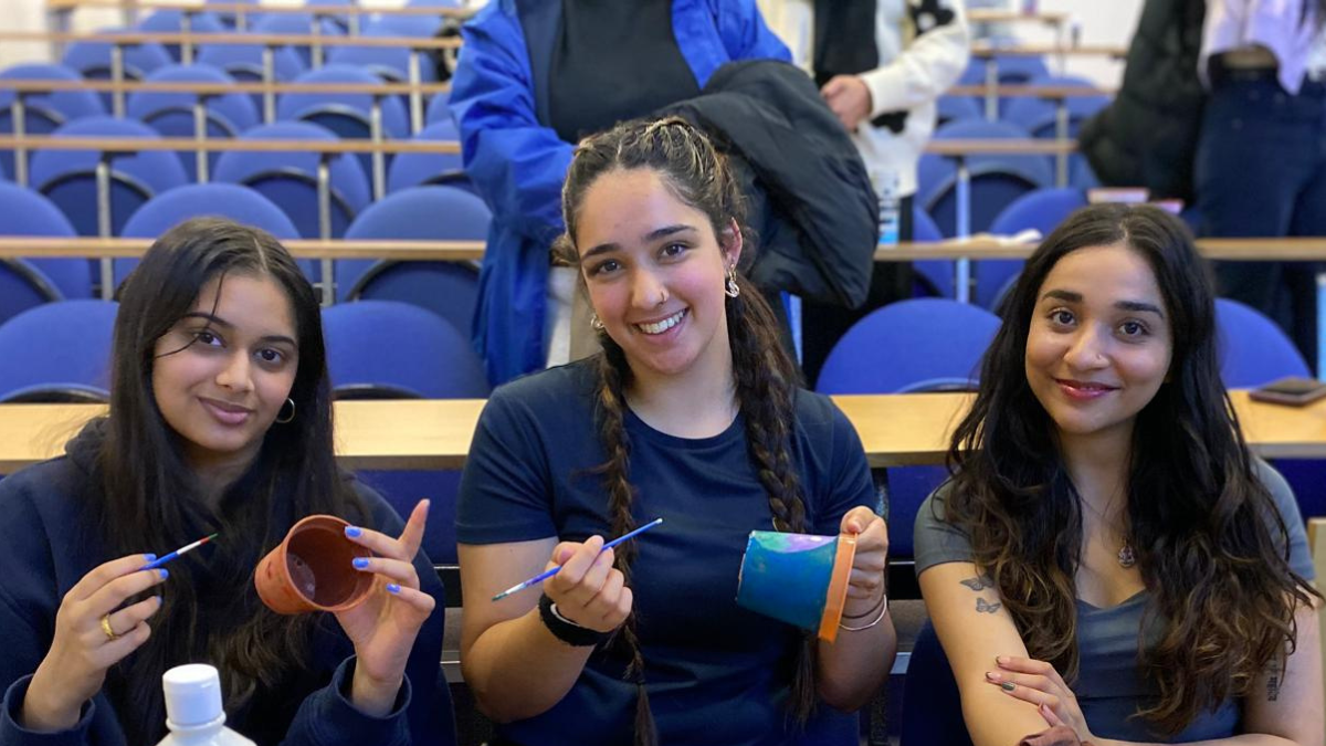 Three people sit in a lecture theatre. They're smiling and holding plant pots they are painting.