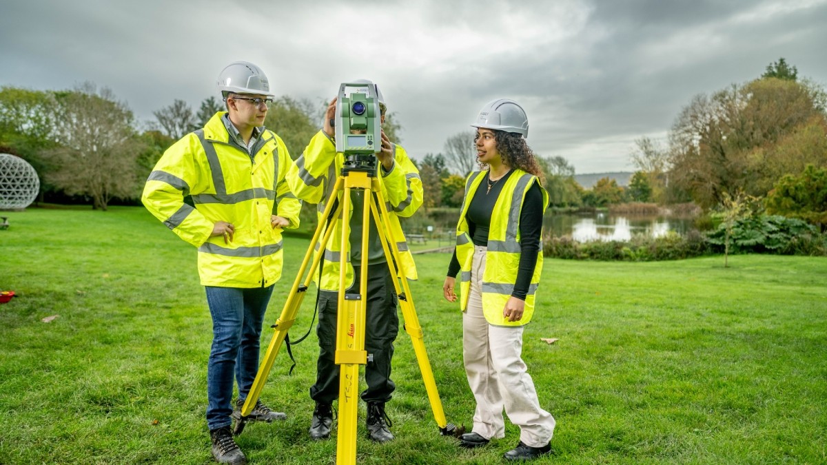  3 students wearing yellow vests using surveying equipment by the University lake