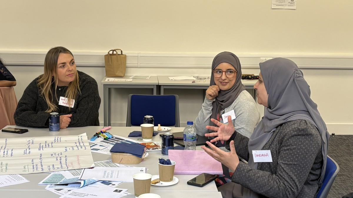 Three women sit at a table talking, with papers laid out in front of them.