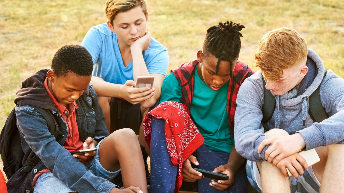 A g roup of four teenage boys sitting on grass looking at mobile phones