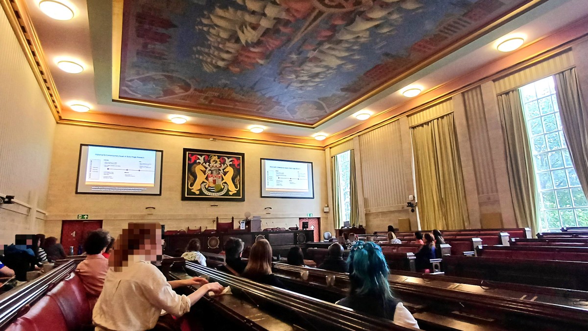 People sit in rows in a large, ornate room with projectors on the wall.