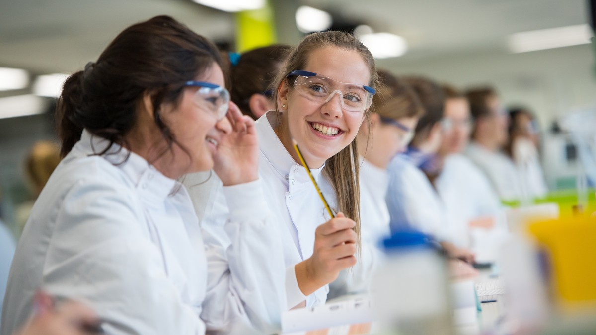 A student in a busy lab with PPE on, smiling at something 