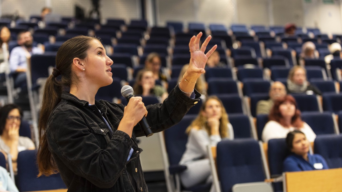 A lecturer talks with a microphone to a room of students. 